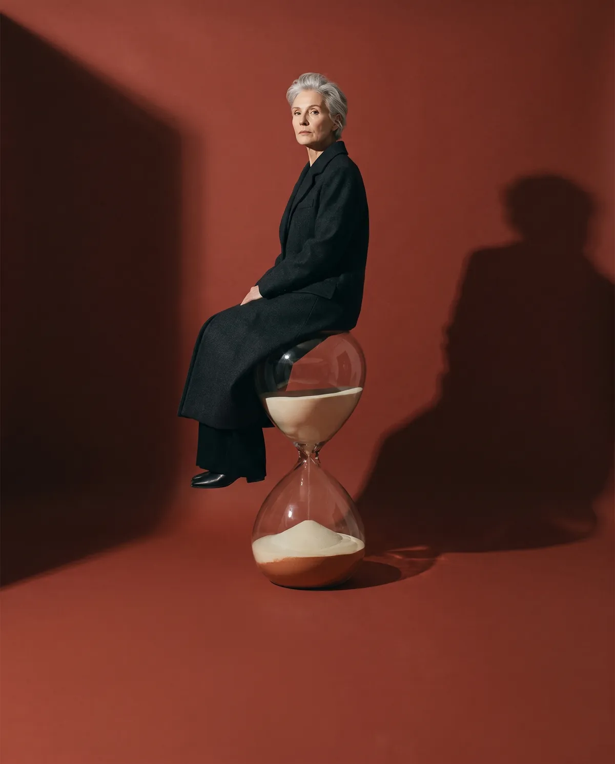 A woman in her sixties in a long black coat sits on a giant hourglass against a rust-red wall.